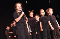 Dziewczynka na scenie śpiewa do mikrofonu, reszta artystów dziecięcych podczas występu w czarnych strojach.