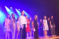 Śpiewający młodzi aktorzy na scenie MOK podczas wykonywania utworu. Widoczne barwne oświetlenie przedstawienia.